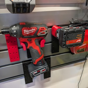 Puede incluir: Un destornillador de instalaci&oacute;n Milwaukee M12 FUEL rojo y negro con una broca amarilla, montado en una pared. Tambi&eacute;n se ven un cargador de bater&iacute;a rojo y una bater&iacute;a. Las herramientas se exhiben en una pared gris con soportes de montaje negros.