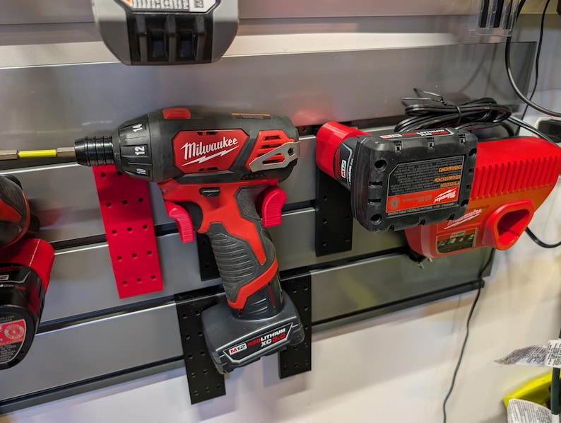 Puede incluir: Un destornillador de instalaci&oacute;n Milwaukee M12 FUEL rojo y negro con una broca amarilla, montado en una pared. Tambi&eacute;n se ven un cargador de bater&iacute;a rojo y una bater&iacute;a. Las herramientas se exhiben en una pared gris con soportes de montaje negros.