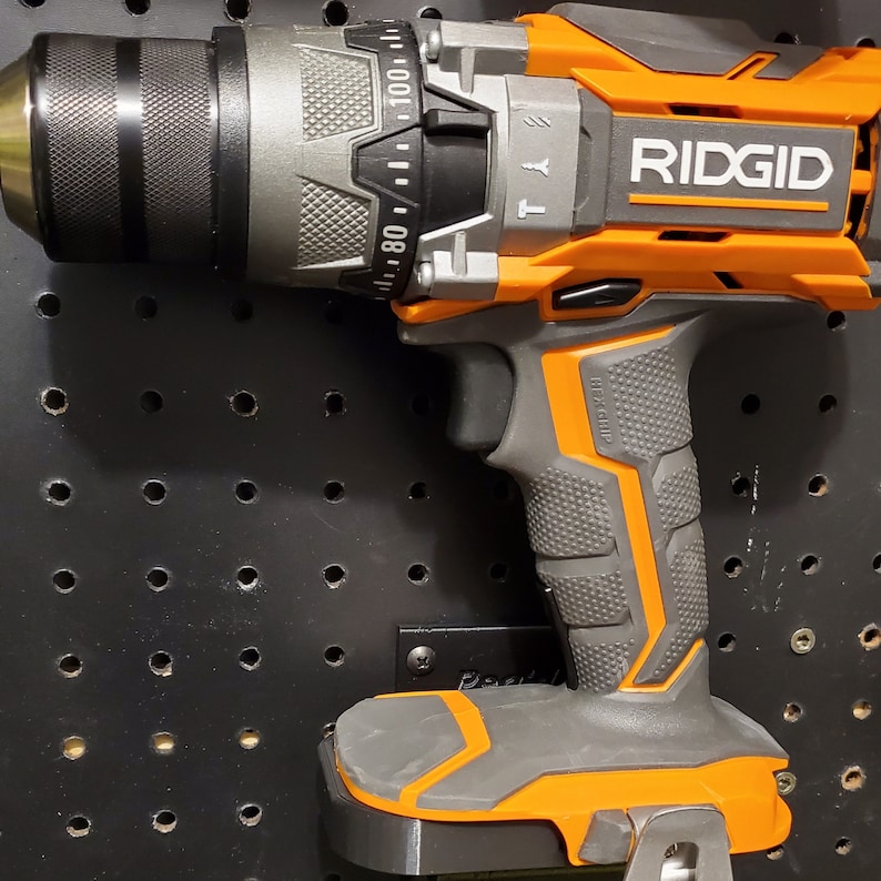 Puede incluir: Un taladro inal&aacute;mbrico negro y naranja con un mango negro y gris. El taladro est&aacute; montado en una placa de pared perforada negra con agujeros. El taladro tiene un portabrocas negro y un gatillo negro y naranja. El taladro tiene un clip de cintur&oacute;n negro y naranja. El taladro tiene un bot&oacute;n de encendido negro y naranja. El taladro tiene un medidor de profundidad negro y naranja. El taladro tiene un dial de control de velocidad negro y naranja. El taladro tiene un dial de control de torque negro y naranja. El taladro tiene una bater&iacute;a negra y naranja. El taladro tiene un cargador negro y naranja. El taladro tiene un estuche de transporte negro y naranja. El taladro tiene un manual negro y naranja. El taladro tiene una tarjeta de garant&iacute;a negra y naranja. El taladro tiene una etiqueta negra y naranja que dice "RIDGID".