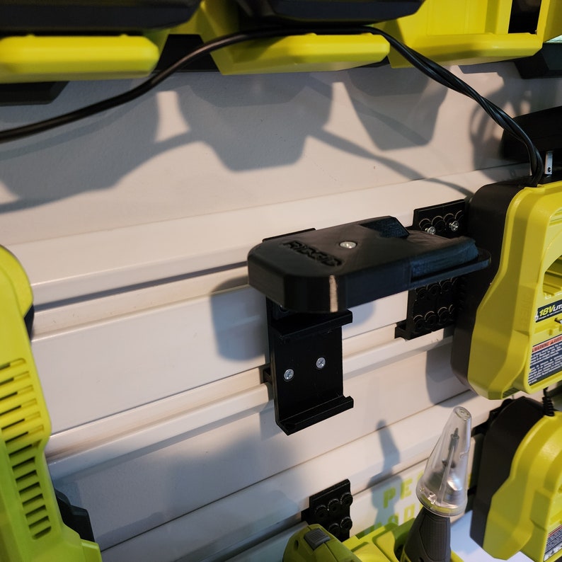 Puede incluir: Un soporte de herramientas de pl&aacute;stico negro con dos tornillos, montado en una pared blanca. El soporte est&aacute; dise&ntilde;ado para sujetar herramientas con mango, como destornilladores o llaves.