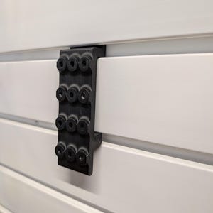 Puede incluir: Un soporte negro con m&uacute;ltiples orificios circulares est&aacute; fijado a una superficie de listones blancos. El soporte parece estar hecho de un material duradero y est&aacute; dise&ntilde;ado para sujetar o soportar algo. La superficie blanca tiene l&iacute;neas horizontales.