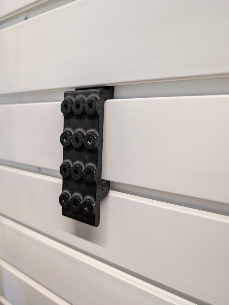 Puede incluir: Un soporte negro con m&uacute;ltiples orificios circulares est&aacute; fijado a una superficie de listones blancos. El soporte parece estar hecho de un material duradero y est&aacute; dise&ntilde;ado para sujetar o soportar algo. La superficie blanca tiene l&iacute;neas horizontales.