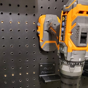 Puede incluir: Un soporte de pared negro Peg'd Products para un taladro con un taladro inal&aacute;mbrico negro y naranja conectado. El taladro tiene una bater&iacute;a negra y naranja y el texto "RIGID" en el lateral.