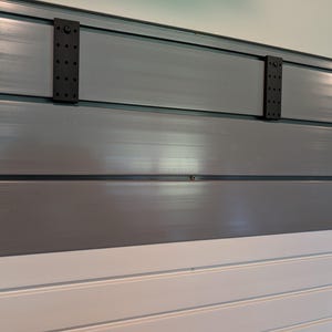May include: A wall-mounted slatwall panel system featuring horizontal gray and white slats. Two rectangular black mounting brackets with multiple circular perforations are attached to the upper gray section of the panel, secured with visible screws.