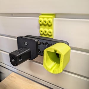 Puede incluir: Un sistema de almacenamiento montado en la pared con un dise&ntilde;o impreso en 3D en negro y amarillo. El sistema incluye un soporte negro con texto y un soporte amarillo. Un soporte amarillo est&aacute; montado encima de los soportes.