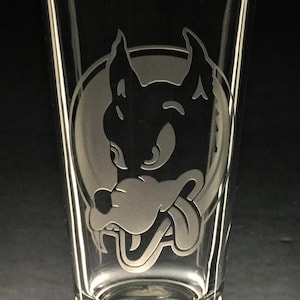May include: A clear glass pint glass with a white etched design of a cartoon wolf's head with its tongue sticking out.
