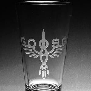 May include: Clear pint glass with an etched design. The design features a stylized bird or winged creature with the word "GODS" above the wings. The glass is transparent and appears to be empty.