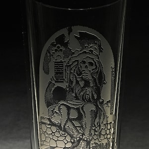 May include: A clear glass pint glass with an etched design of a skeleton figure wearing a hooded robe. The skeleton is sitting on a throne with a crown on its head.