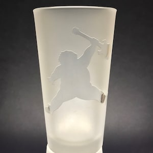 Puede incluir: Un vaso esmerilado con una silueta blanca de una persona tocando una guitarra. La persona está saltando en el aire con la guitarra levantada por encima de su cabeza.