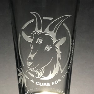 Puede incluir: Un vaso de pinta de vidrio transparente con un diseño grabado en blanco de una cabra con una hoja de marihuana en la boca. El texto "A Cure for the Bleakness" está grabado alrededor de la cabra.