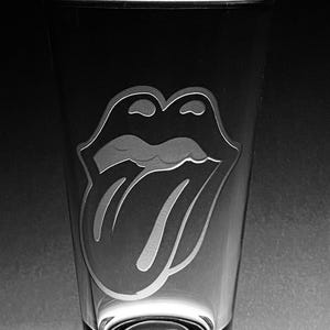 May include: A clear glass tumbler featuring an engraved image of a pair of lips and a tongue, a well-known rock and roll logo. The glass has a cylindrical shape with a flared top and a solid base. The background is dark, creating a contrast with the design.