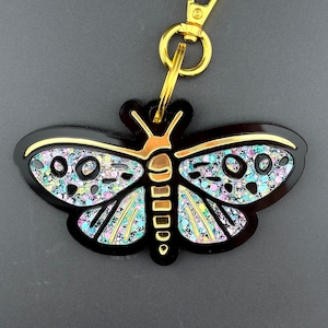 May include: A butterfly-shaped keyring featuring a black acrylic base with gold-toned outlines. The wings are filled with a multi-coloured glitter inlay in shades of blue, pink, and silver. A gold-coloured metal lobster clasp is attached to the top.