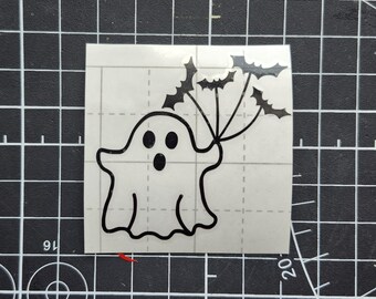 May include: A black and white Halloween-themed sticker featuring a cartoon ghost holding a cluster of bats. The ghost has a surprised expression with large, round eyes and an open mouth. The bats are silhouetted and add a spooky touch to the design.