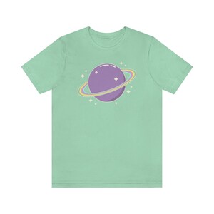 K&ouml;nnte beinhalten: Ein hellgr&uuml;nes T-Shirt mit einem lila Planeten mit einem Regenbogenring darum. Der Planet ist von kleinen wei&szlig;en Sternen umgeben.