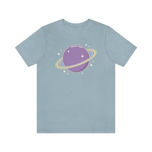 K&ouml;nnte beinhalten: Ein hellblaues T-Shirt mit einem lila Planeten mit einem Regenbogenring darum. Der Planet ist von kleinen wei&szlig;en Sternen umgeben.