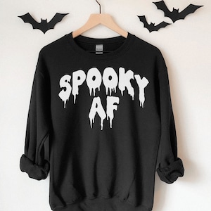 Peut inclure: Sweat-shirt noir avec un motif blanc qui dit "Spooky AF" en lettres qui coulent.