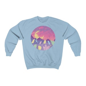May include: Light blue sweatshirt with a circular graphic of a crescent moon, mountains, and a pink and purple sunset. The graphic is surrounded by a ring of pastel-colored crystals.