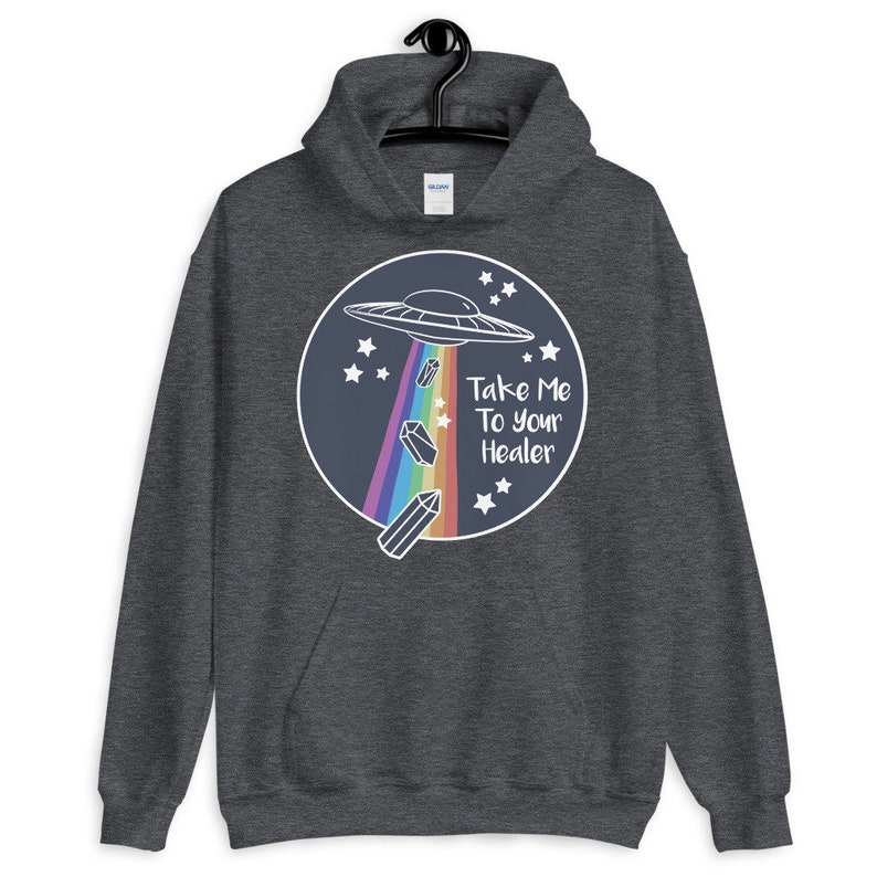 May include: A dark grey hoodie with a graphic of a UFO flying over a rainbow with crystals falling from it. The text "Take Me To Your Healer" is written in white on a blue circle.