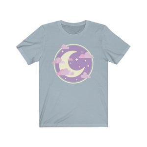May include: A light blue t-shirt with a purple circle design featuring a crescent moon and pink clouds. The moon is yellow and there are small white stars inside the circle.