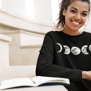 Puede incluir: Sudadera negra de manga larga con un diseño de fases lunares en blanco y gris. La persona lleva la sudadera al aire libre con un libro y un cuaderno.