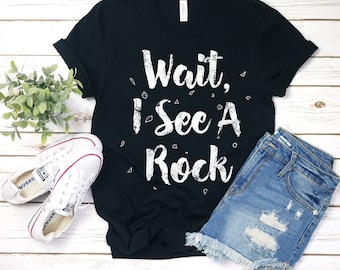Puede incluir: Camiseta negra con texto blanco que dice "Wait, I See A Rock". La camiseta está sobre una superficie de madera blanca con un par de zapatillas blancas, una planta verde y unos pantalones cortos de mezclilla azul.