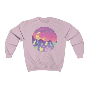May include: A pink sweatshirt with a graphic of a crescent moon, mountains, and crystals in a circle against a sunset background.