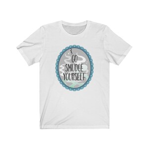 以下が含まれることがあります： 白いTシャツに、青とグレーの円形のデザインが施され、黒い文字で「GO SMUDGE YOURSELF」と書かれています。