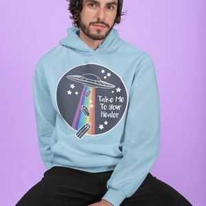May include: A light blue hoodie with a black circle graphic featuring a UFO, rainbow, crystals, and the text "Take Me To Your Healer".