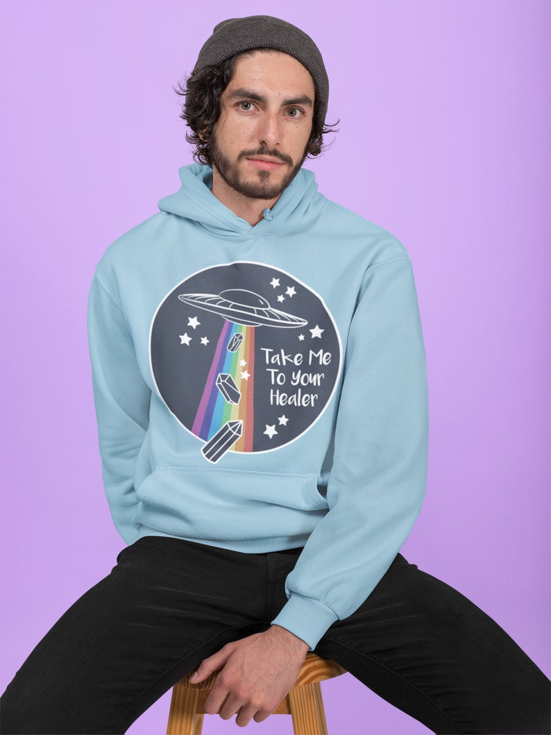 May include: A light blue hoodie with a black circle graphic featuring a UFO, rainbow, crystals, and the text "Take Me To Your Healer".