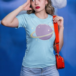 K&ouml;nnte beinhalten: Eine Frau tr&auml;gt ein hellblaues T-Shirt mit einem lila Planeten und einem Regenbogenring-Motiv. Sie tr&auml;gt auch blaue Jeans und eine rote Umh&auml;ngetasche.