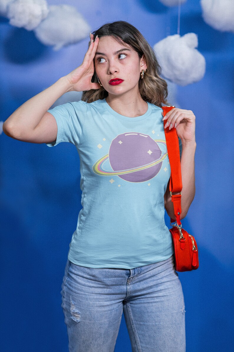 K&ouml;nnte beinhalten: Eine Frau tr&auml;gt ein hellblaues T-Shirt mit einem lila Planeten und einem Regenbogenring-Motiv. Sie tr&auml;gt auch blaue Jeans und eine rote Umh&auml;ngetasche.