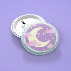 Puede incluir: Un botón azul claro con una luna creciente y nubes rosadas sobre un fondo morado. El botón tiene un pasador plateado en la parte posterior.