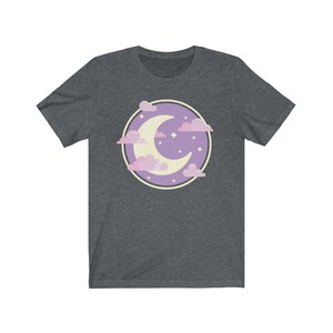 May include: A dark heather grey t-shirt with a purple circle design featuring a crescent moon and pink clouds. The moon is yellow and there are small white stars in the circle.