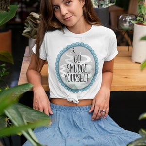 以下が含まれることがあります： 「Go Smudge Yourself」と書かれた青と灰色の円形のグラフィックが描かれた白いTシャツ。長い茶色の髪と青いスカートの女性が着用しています。