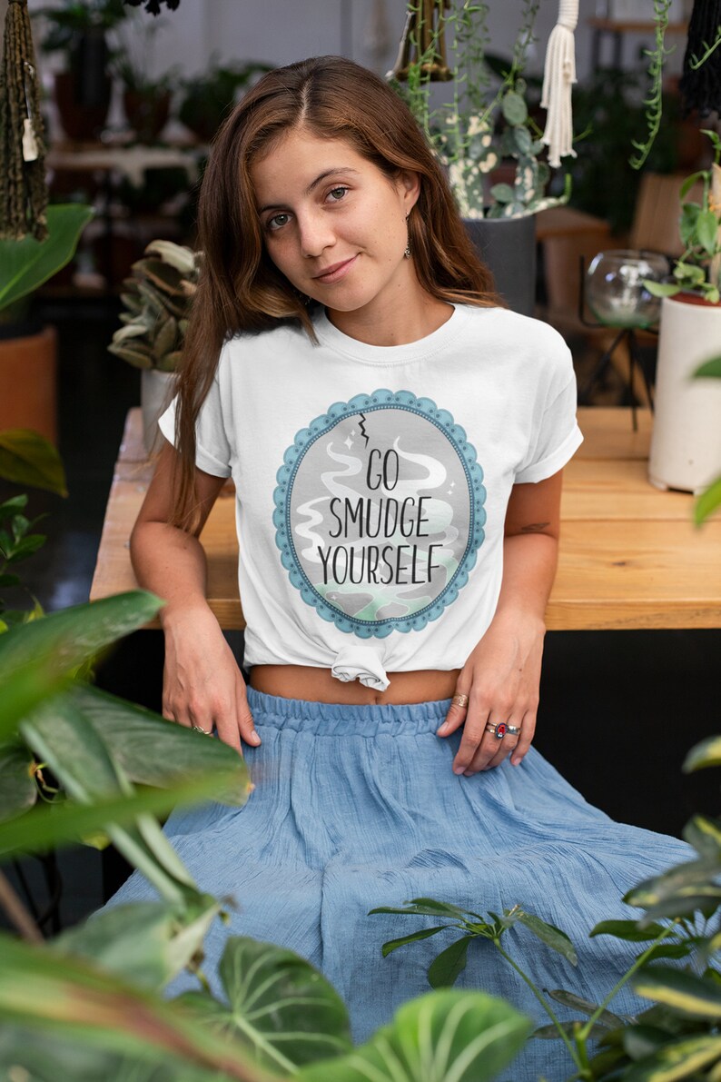 以下が含まれることがあります： 「Go Smudge Yourself」と書かれた青と灰色の円形のグラフィックが描かれた白いTシャツ。長い茶色の髪と青いスカートの女性が着用しています。