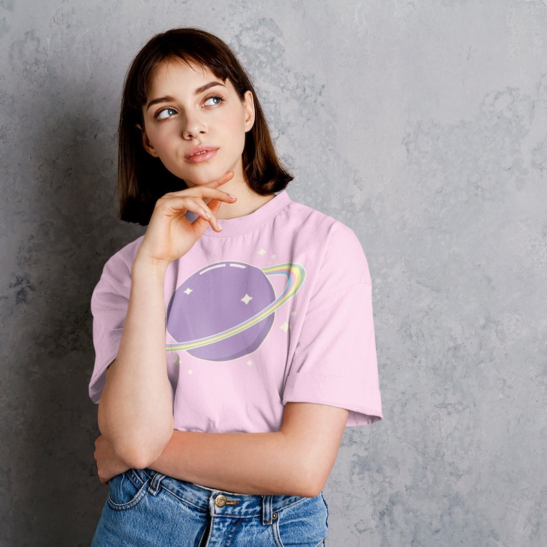 K&ouml;nnte beinhalten: Eine Frau tr&auml;gt ein pinkfarbenes T-Shirt mit einem lila Planeten und einem Regenbogenring-Motiv. Das Shirt hat einen Rundhalsausschnitt und kurze &Auml;rmel. Sie tr&auml;gt eine blaue Jeans und hat kurze braune Haare.