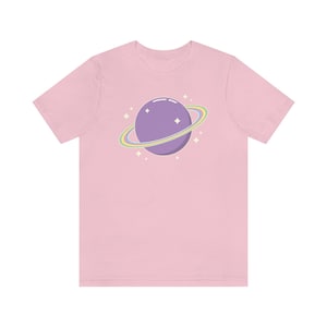 K&ouml;nnte beinhalten: Ein pinkfarbenes T-Shirt mit einem lila Planeten mit einem Regenbogenring und wei&szlig;en Sternen.
