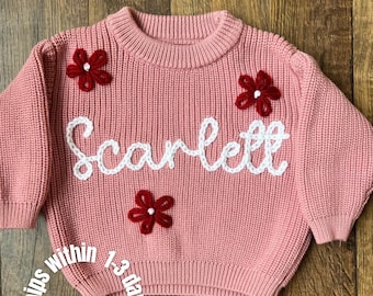 May include: A dusty pink long-sleeve knit sweater featuring a crew neckline and ribbed cuffs. The front is decorated with three dark red yarn flowers with white centers and the name Scarlett embroidered in white cursive yarn lettering.