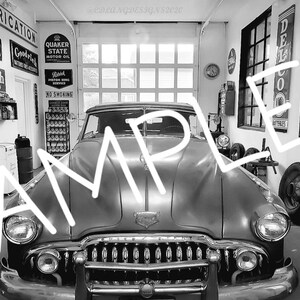 May include: A black and white photo of a vintage car in a garage. The car is a 1940s Buick Super with a chrome grille and headlights. The garage is filled with old signs and tools.