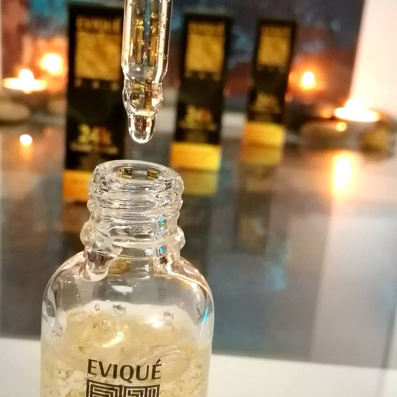May include: A clear glass bottle with a gold liquid and gold flakes inside. The bottle is labeled "EVIQUE" and has a gold design. A dropper is dispensing a drop of the liquid.