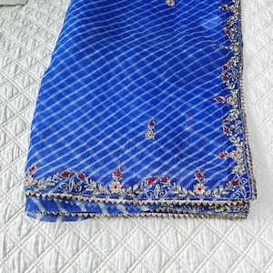 May include: A folded, sheer blue fabric with a white striped pattern. The fabric is embellished with silver and red floral embroidery along the edges. The fabric is likely a scarf or shawl.