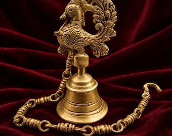 May include: A brass bell featuring a detailed peacock handle with intricate engraved patterns. The bell is attached to a textured metal chain with a hook at one end, showcasing a metallic gold finish and a classic flared bell shape design.