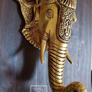 Puede incluir: Un llamador de puerta de metal dorado con forma de cabeza de elefante con detalles intrincados. La trompa del elefante est&aacute; enroscada en forma de anillo.