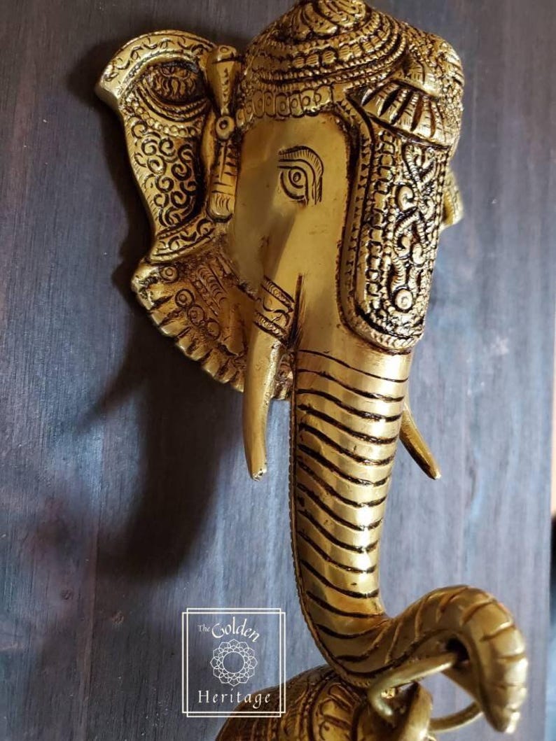 Puede incluir: Un llamador de puerta de metal dorado con forma de cabeza de elefante con detalles intrincados. La trompa del elefante est&aacute; enroscada en forma de anillo.