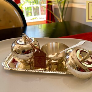 Puede incluir: Una bandeja plateada con tres recipientes plateados en forma de manzana. La bandeja tiene un borde con cuentas. Los recipientes tienen tapas y uno tiene una peque&ntilde;a cuchara. La bandeja tiene una etiqueta que dice "The Golden Heritage".