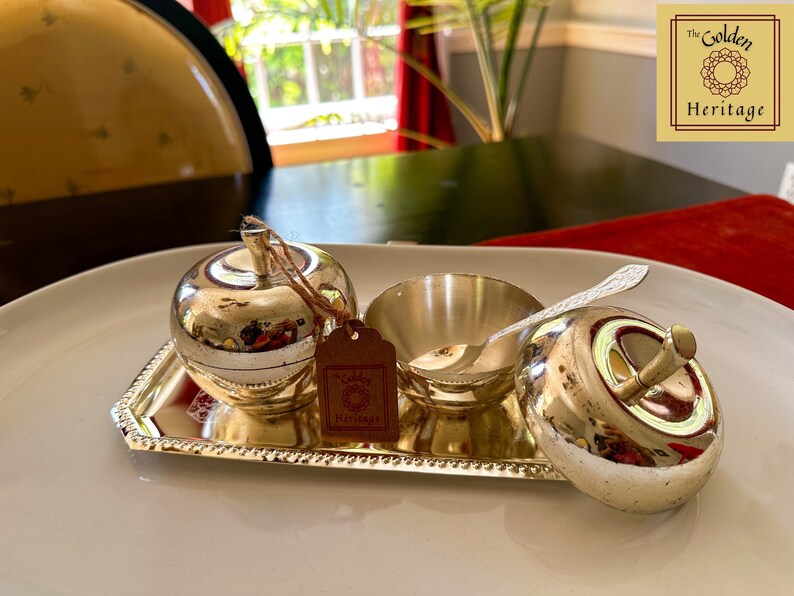 Puede incluir: Una bandeja plateada con tres recipientes plateados en forma de manzana. La bandeja tiene un borde con cuentas. Los recipientes tienen tapas y uno tiene una peque&ntilde;a cuchara. La bandeja tiene una etiqueta que dice "The Golden Heritage".