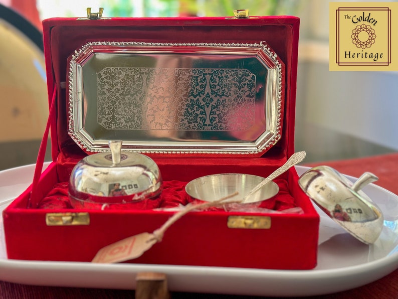 Puede incluir: Un juego de servicio de plata en una caja de terciopelo rojo. El juego incluye dos cuencos con forma de manzana, un cuenco peque&ntilde;o y una cuchara. La caja tiene una bandeja de plata con un dise&ntilde;o grabado. La caja tiene una etiqueta que dice "The Golden Heritage".