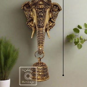 Puede incluir: Colgante de pared de metal dorado en forma de cabeza de elefante con detalles intrincados y una campana colgando del tronco. La cabeza de elefante mide 10,5 pulgadas de alto y 4 pulgadas de ancho.