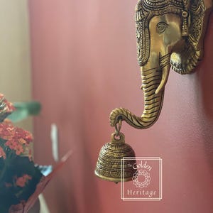 Puede incluir: Una decoraci&oacute;n de pared de metal dorado en forma de cabeza de elefante con detalles intrincados. La trompa del elefante est&aacute; enroscada y sostiene una peque&ntilde;a campana adornada. La decoraci&oacute;n de pared est&aacute; etiquetada como "The Golden Heritage".