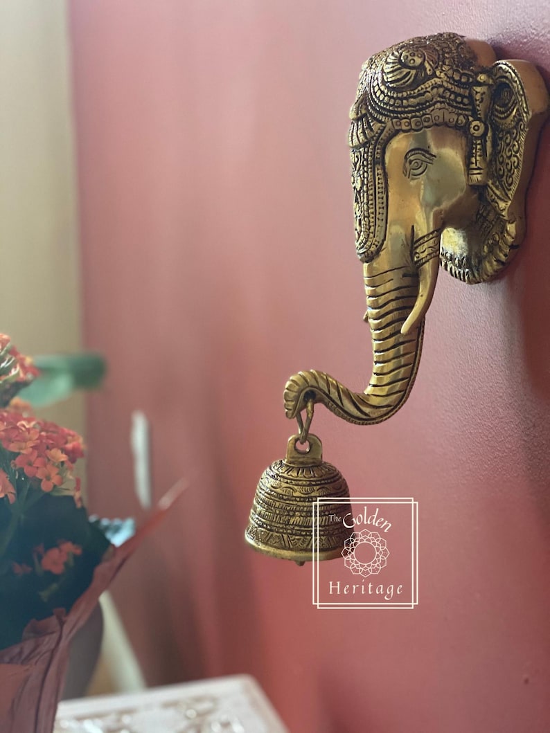 Puede incluir: Una decoraci&oacute;n de pared de metal dorado en forma de cabeza de elefante con detalles intrincados. La trompa del elefante est&aacute; enroscada y sostiene una peque&ntilde;a campana adornada. La decoraci&oacute;n de pared est&aacute; etiquetada como "The Golden Heritage".